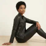 Woman wearing black leather moto jacket seated on floor, styled with black leather pants and hoop earrings for complete edgy-chic look