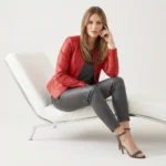 Women's red leather bomber jacket with V-neckline, front zipper closure, ribbed cuffs and hem, styled on white modern chair - front view