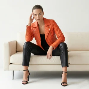 Woman wearing vibrant orange quilted leather moto jacket - Urban Dynasty Leather Jacket - with asymmetric zipper and diamond stitching sitting on beige sofa